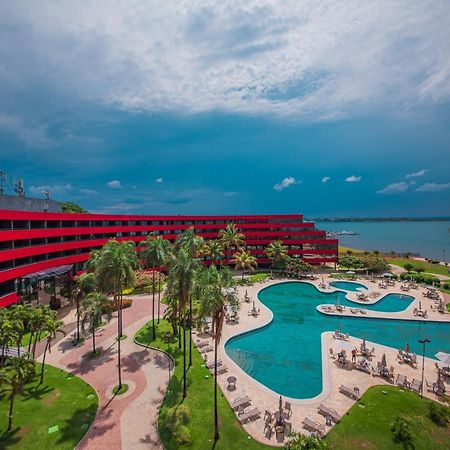 فندق رويال توليب برازيليا ألفورادا المظهر الخارجي الصورة The photo shows a modern hotel building with a striking red exterior, surrounded by lush greenery. In the foreground, there is a large, creatively shaped swimming pool, with lounge chairs arranged around it. The backdrop features a body of water, pos