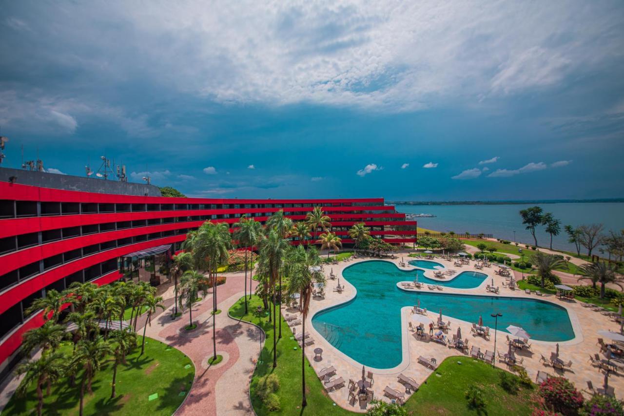 فندق رويال توليب برازيليا ألفورادا المظهر الخارجي الصورة The photo shows a modern hotel building with a striking red exterior, surrounded by lush greenery. In the foreground, there is a large, creatively shaped swimming pool, with lounge chairs arranged around it. The backdrop features a body of water, pos