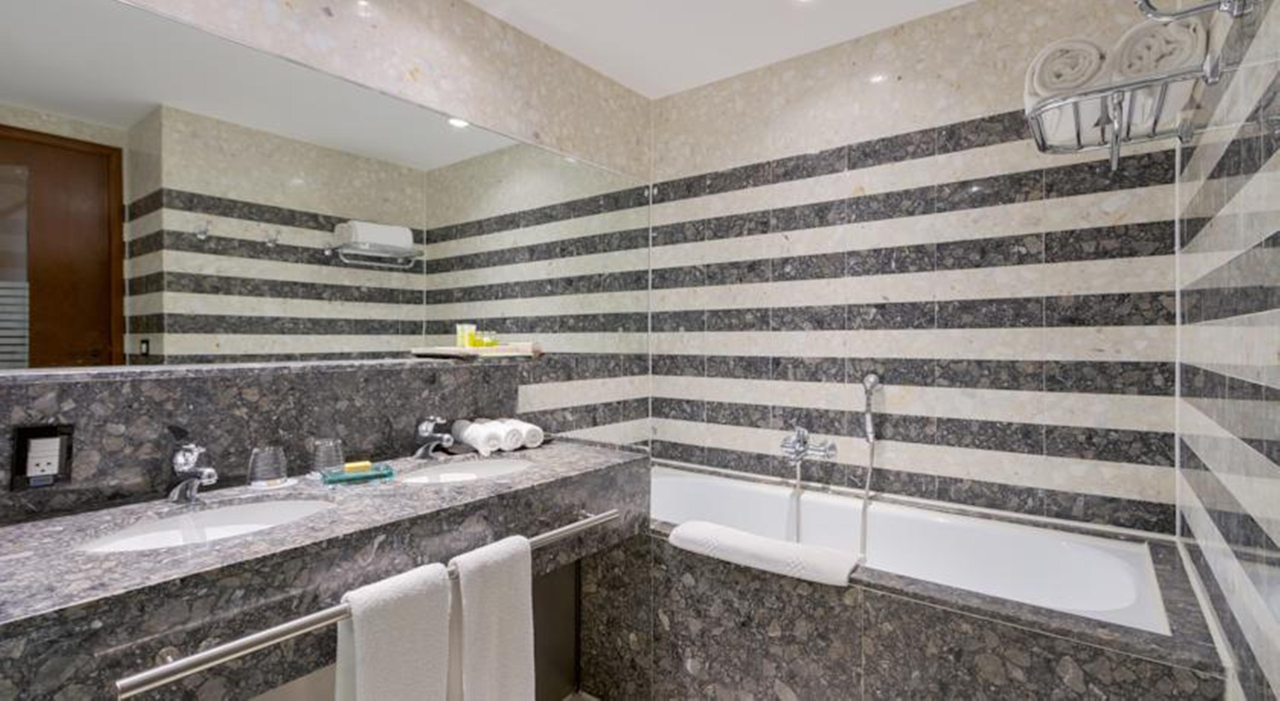 فندق رويال توليب برازيليا ألفورادا المظهر الخارجي الصورة The photo shows a modern bathroom featuring a combination of marble and stone finishes. The walls are decorated with horizontal stripes in various shades of gray and cream. There is a bathtub with a white exterior, and a showerhead is mounted on the 