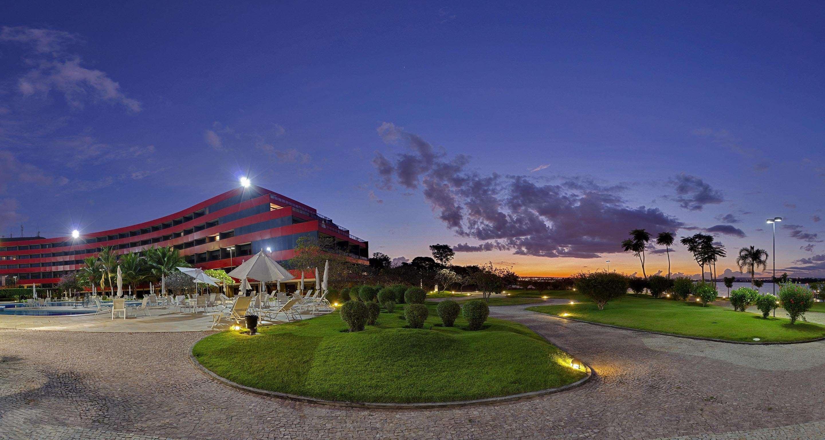فندق رويال توليب برازيليا ألفورادا المظهر الخارجي الصورة The photo depicts a modern building, possibly a hotel or resort, with a striking red exterior. It is set in a landscaped area that features well-maintained greenery and ornamental plants. In the foreground, there are lounge areas with umbrellas, sugg