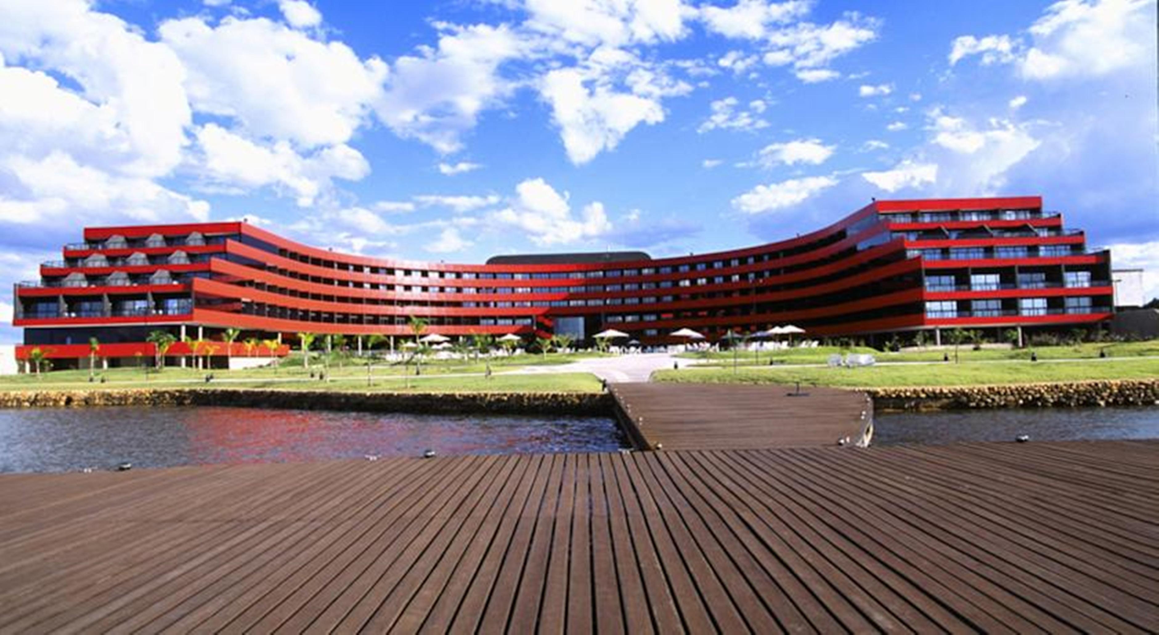 فندق رويال توليب برازيليا ألفورادا المظهر الخارجي الصورة The image shows a modern, curved building with a striking red facade, situated near a body of water. The architecture features multiple levels with large windows that create a sleek and contemporary appearance. In front of the building, there is a wo