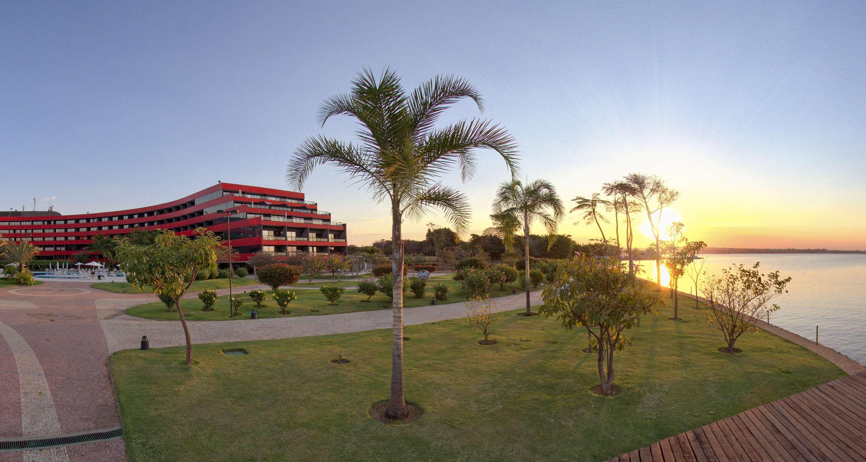 فندق رويال توليب برازيليا ألفورادا المظهر الخارجي الصورة The photo depicts a scenic view featuring a red multi-story building alongside a serene waterfront. In the foreground, there are several palm trees and lush green lawns, creating a tropical atmosphere. The sun is setting in the background, casting wa