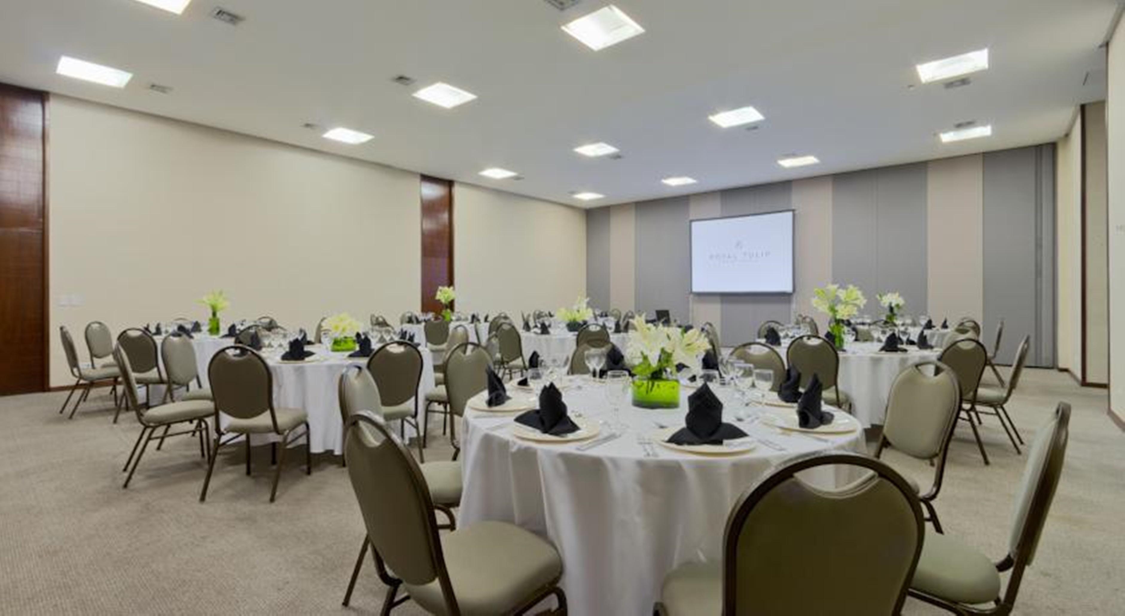 فندق رويال توليب برازيليا ألفورادا المظهر الخارجي الصورة The photo shows a banquet or conference room set up with multiple round tables covered in white tablecloths. Each table is arranged with black napkins and centerpieces featuring flowers, likely in shades of green and white. The room has beige walls a