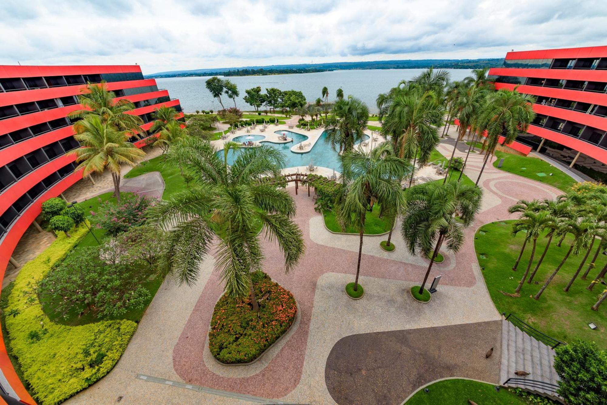 فندق رويال توليب برازيليا ألفورادا المظهر الخارجي الصورة The photo shows a scenic view of a courtyard area in a residential complex. There are multiple red buildings surrounding a pool area, which is surrounded by palm trees and landscaped gardens. The pool features a curvy design, and there are pathways m